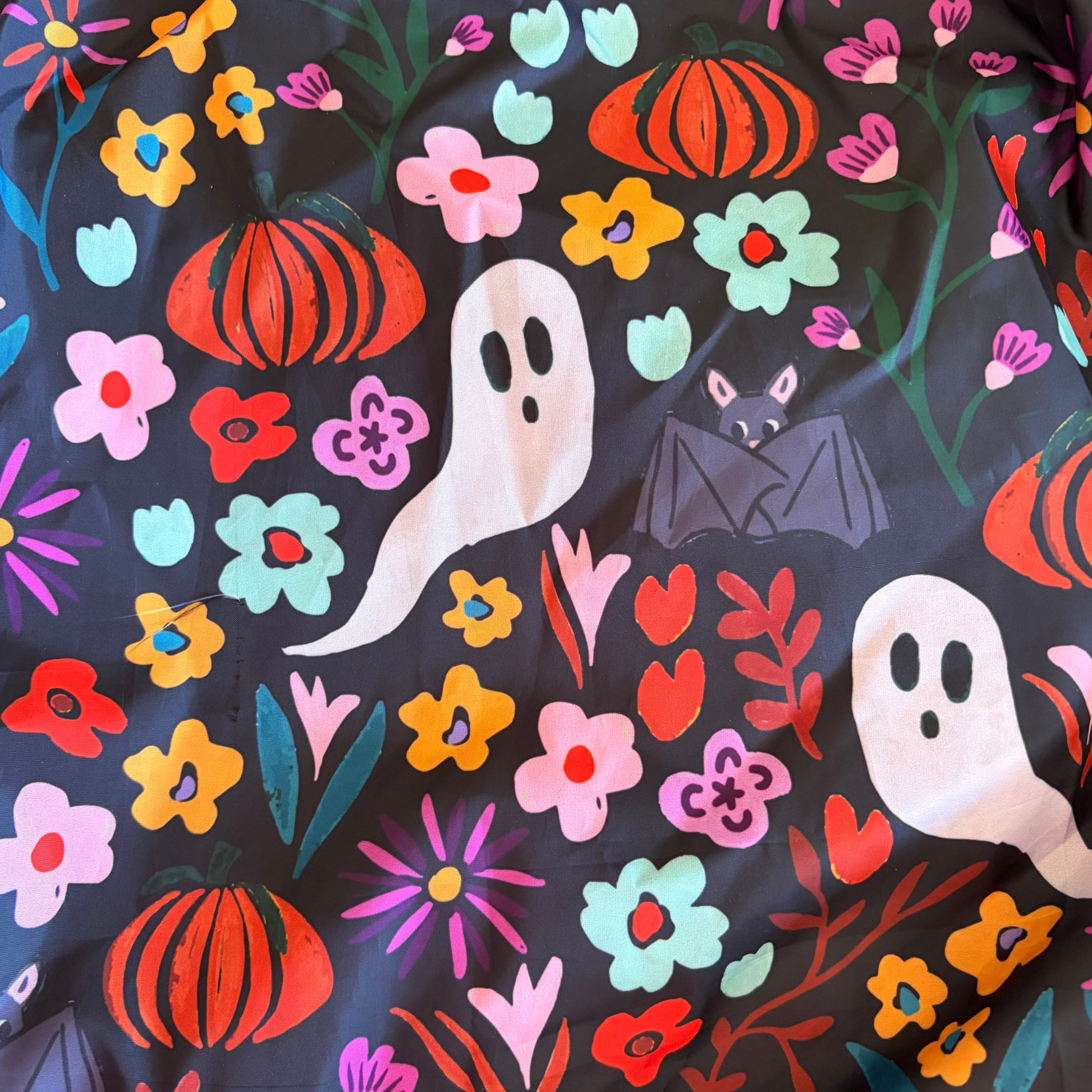 A close-up of a Halloween-themed sensory parachute design featuring orange pumpkins, white ghosts, pink and orange bats, and colourful flowers on a black background.